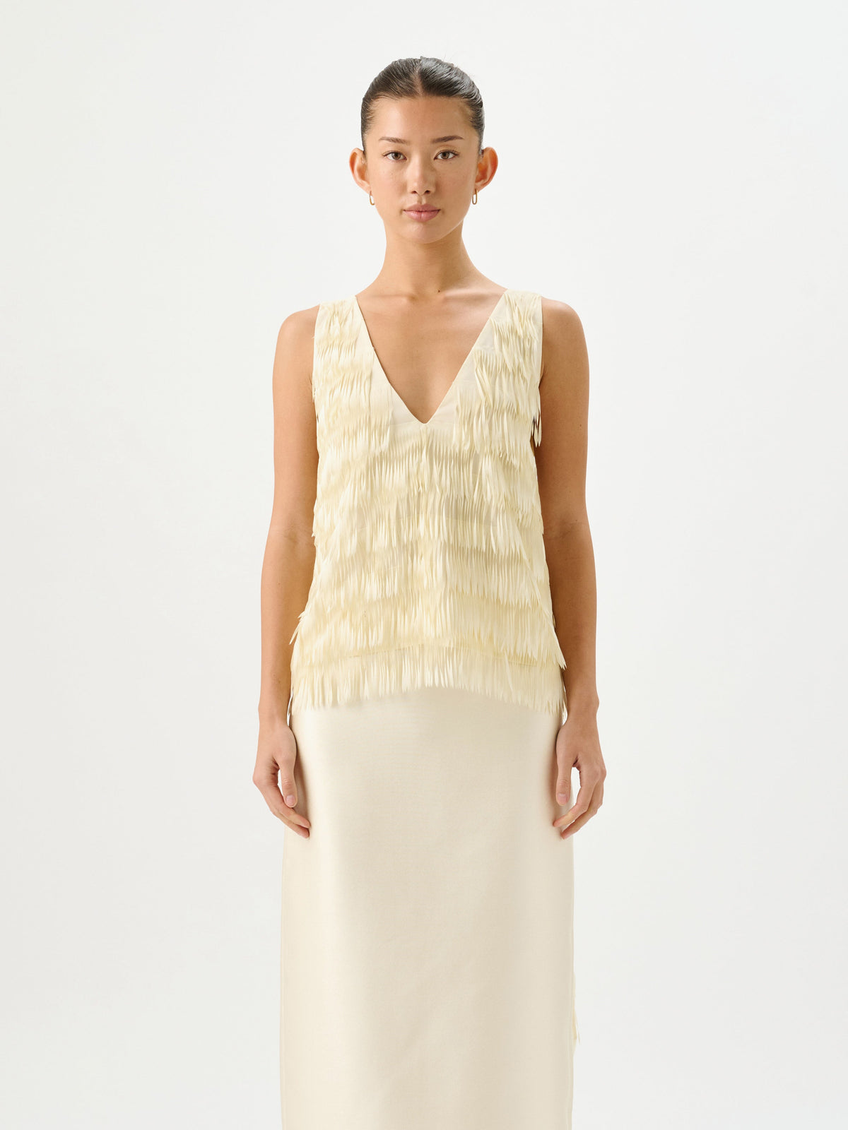 A model stands against a plain white studio background wearing a sleeveless cream dress with a textured, ruffled V-neck bodice and a long, smooth skirt.