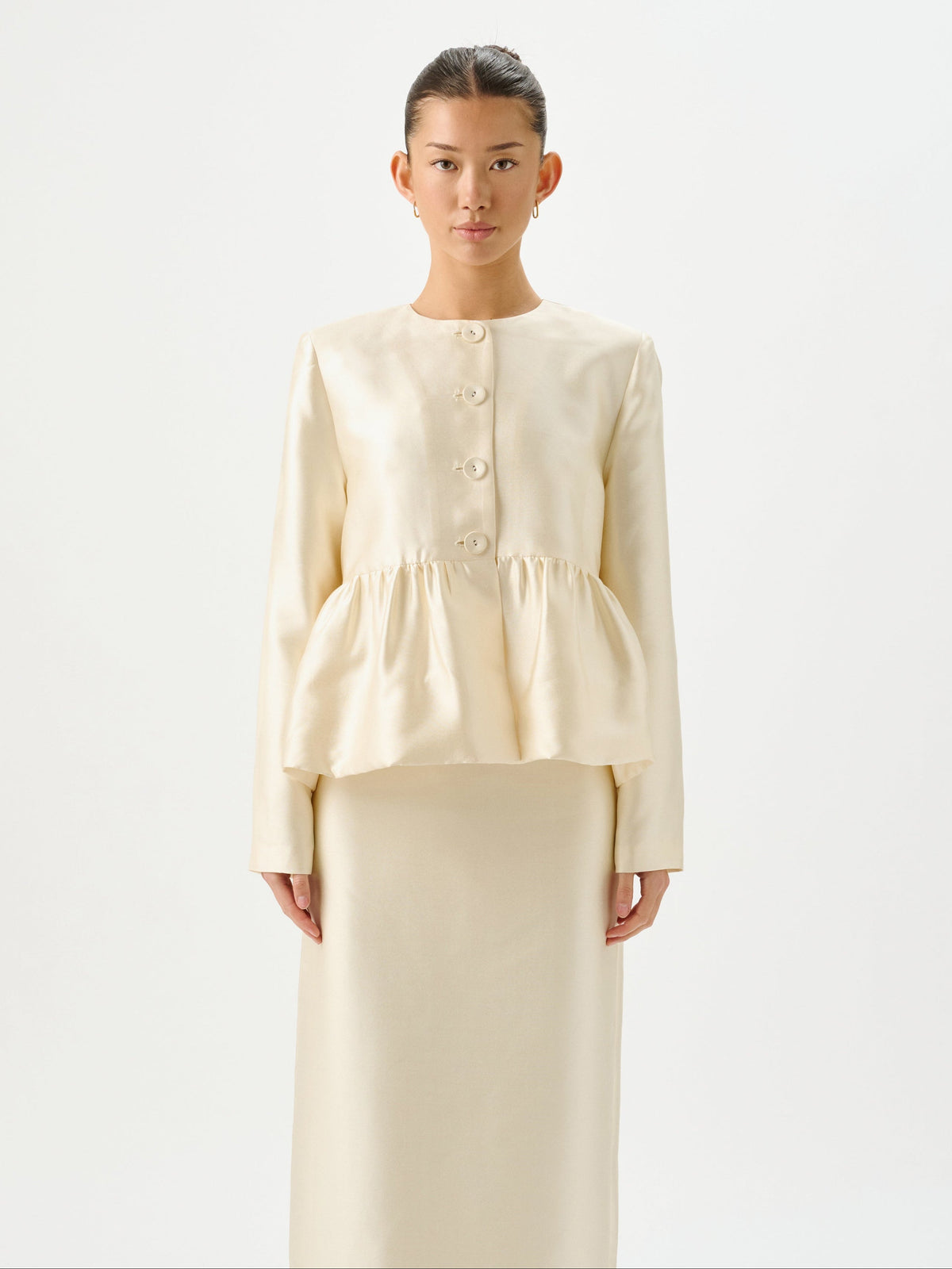A model stands against a plain white studio background wearing a matching cream-colored two-piece outfit consisting of a long-sleeve peplum-style top with decorative buttons and a long skirt.